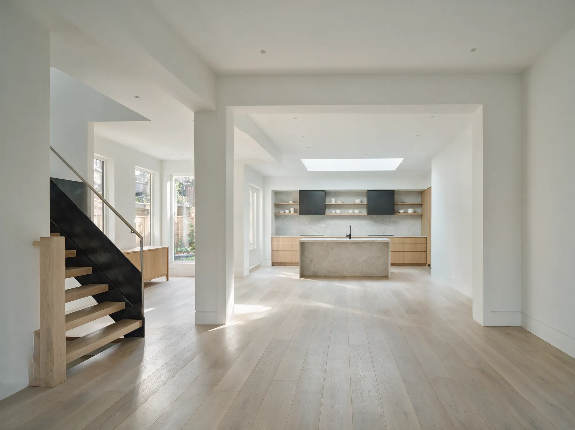 An open main floor after a whole-home renovation, with oak floors, a steel-and-oak staircase and a stone island under a skylight